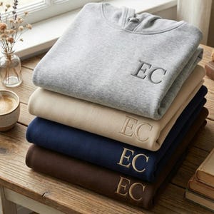 May include: A stack of five folded sweatshirts in various colors: gray, beige, navy blue, and brown. Each sweatshirt has the embroidered initials "EC" on the front. The sweatshirts are neatly arranged on a wooden surface.