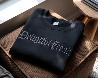 May include: Black crew neck sweatshirt featuring the text "Delightful Freak" in a gothic font. The sweatshirt is folded on a brown cushion, with a wooden side table and a window in the background.