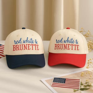 May include: Two baseball caps with the text "red, white & BRUNETTE". One cap has a navy blue brim, the other a red brim. Both caps have a cream-colored crown. An American flag is in the foreground.