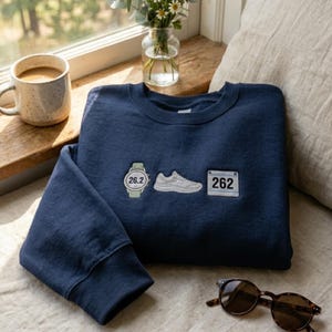 May include: Navy blue sweatshirt with embroidered patches of a watch showing 26.2, a running shoe, and a race bib with the number 262. A mug of coffee, sunglasses, and a vase of flowers are also in the image.