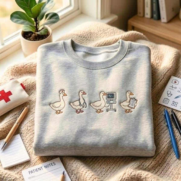 May include: A light grey crewneck sweatshirt featuring an embroidered graphic of four white geese dressed as medical professionals, including one with a nurse cap, one with a stethoscope, one with a heart monitor, and one holding a clipboard.