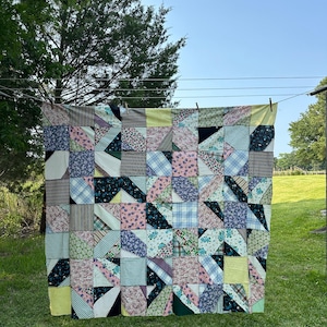 Op de afbeelding: Een kleurrijke patchworkquilt met verschillende stofpatronen, waaronder bloemen-, ruit- en streepontwerpen. De quilt wordt buiten op een waslijn getoond. De quilt meet ongeveer 178 cm bij 203 cm. De quilt is gemaakt van verschillende kleuren.
