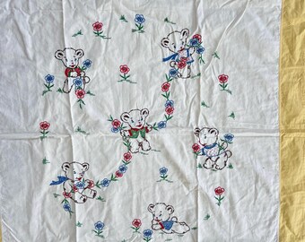 Op de afbeelding: Witte babydeken met geborduurde teddyberen met bloemen. De beren dragen rode, blauwe en groene accenten. De deken heeft een gele rand.