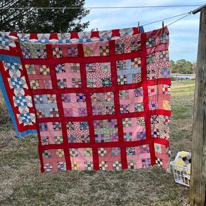 Puede incluir: Una colcha de retazos roja, rosa y azul colgada de un tendedero al aire libre. La colcha está hecha de muchos cuadrados pequeños cosidos juntos y tiene un borde rojo.