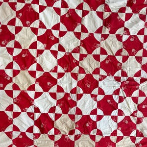 May include: A handmade quilt with a repeating pattern of red and white shapes. The red sections feature a floral design. The quilt's design is a mix of curved and square shapes, creating a visually appealing textile.