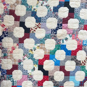 May include: A patchwork quilt with a repeating pattern of off-white octagons and colorful squares. The squares feature floral, polka dot, and plaid designs in shades of red, blue, green, and pink. The quilt has a vintage, handmade aesthetic.