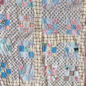 Op de afbeelding: Een patchworkquilt met verschillende patronen in blauw, roze en bruin. De quilt heeft een geruit patroon en een raster van crèmekleurige stof met rode lijnen. De vierkanten zijn gemaakt van bloemen- en geometrische stoffen.