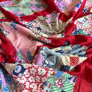 Op de afbeelding: Een vintage patchworkquilt met een levendige mix van bloemen, geometrische en effen stoffen. De quilt heeft een heldere rode rand en achterkant, met een verscheidenheid aan kleuren, waaronder roze, blauw, groen en wit. Handgemaakt textiel.