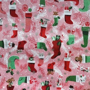 May include: Pink fabric with a repeating pattern of Christmas stockings in red and green, filled with gifts, candy canes, and small toys. The design includes white and silver accents, creating a festive holiday theme.