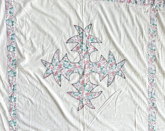 Op de afbeelding: Crèmekleurige quilt met een centraal bloemenontwerp en bloemenrand. Het centrale ontwerp is een gestileerde bloemvorm gevuld met een roze, groen en blauw bloemenpatroon. De rand is een herhalend bloemenpatroon.