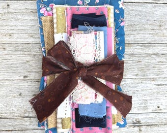 Könnte beinhalten: Ein Stoffbündel mit einer Vielzahl von Texturen und Farben, darunter blau floral, rosa und Jute. Das Bündel ist mit einem braunen Band mit einem Tupfenmuster gebunden. Das Bündel enthält Spitze und Stoffreste.