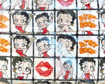 Op de afbeelding: Een witte stof met een herhalend patroon van Betty Boop-illustraties in zwart, rood en goud. Het ontwerp bevat Betty Boops gezicht, rode lippen en de woorden "Betty Boop".