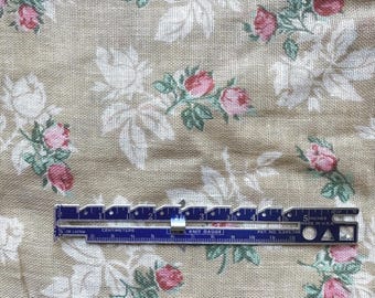 May include: A beige fabric with a floral pattern of pink roses and white leaves. A blue knit gauge, marked in inches and centimeters, lies across the fabric. The gauge reads "KNIT GAUGE" and "MADE IN U.S.A."