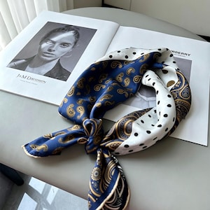 May include: A square scarf featuring a dual-pattern design with a deep blue section adorned with gold paisley motifs and a white section decorated with black polka dots. The scarf is tied in a knot, showcasing the contrasting patterns and colours.