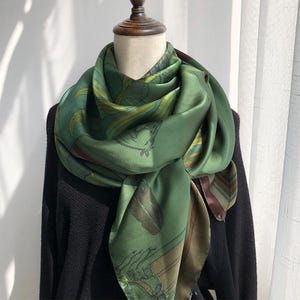 May include: A square silk scarf in shades of olive and forest green featuring a printed pattern of horse heads and foliage. The scarf is draped around the neck of a mannequin wearing a black textured sweater, showcasing a smooth, satin-like finish.