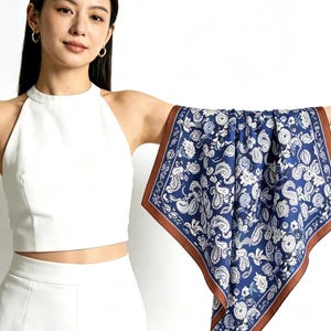 May include: A navy blue scarf with a white paisley pattern and a brown border. The square scarf is displayed, highlighting its design. The scarf is made of a soft, silky material.