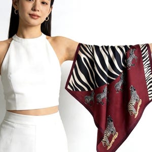 May include: A square scarf with a zebra print. The scarf has a burgundy border and a central panel with illustrations of zebras. The zebra print is black and white. The scarf is held up by a person.