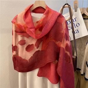 May include: A sheer, red and pink scarf with a cloud-like pattern. The scarf is draped over a white dress and a light pink blouse. The scarf is hanging on a wooden hanger. The background includes a brown purse and a white bag with the text "Bar celo na".