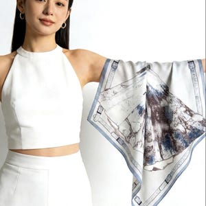 May include: A square silk scarf featuring a landscape design in blue, grey, and brown tones. The scarf has a light blue border with a buckle pattern. The scarf is held up in front of a white top and trousers.