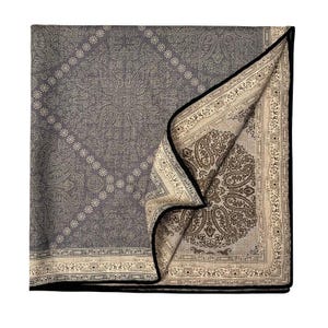 May include: A folded, gray paisley patterned textile with a black border. The textile features a detailed design with a lighter beige color, creating a contrast. The corner is folded over, revealing a different paisley pattern.