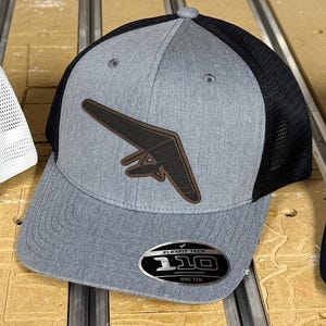 Può includere: Cappellino da baseball Flexfit tech grigio e nero con una toppa di deltaplano in pelle marrone e il testo "110 One Ten" sul davanti.