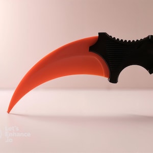 May include: A black and orange karambit knife with a curved blade and a finger ring. The blade is black and the handle is orange.