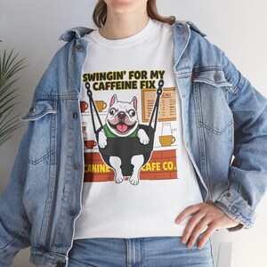 Könnte beinhalten: Weißes T-Shirt mit einem französischen Bulldoggen-Motiv auf einer Schaukel, dem Text "SWINGIN' FOR MY CAFFEINE FIX" und "CANINE CAFE CO." Der Hund trägt ein grünes Halsband. Der Hintergrund zeigt Kaffeetassen und eine Speisekarte.