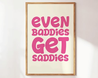 May include: A framed art print with the words "even baddies get saddies" in bright pink, bubble-style letters. The text is centered on a cream-colored background, and the frame is a light brown wood.