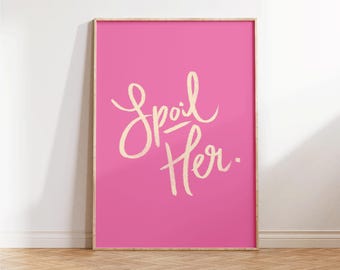 May include: A framed pink art print with the words "Spoil Her" in a cursive font. The artwork is displayed on a wooden floor and leans against a white wall.