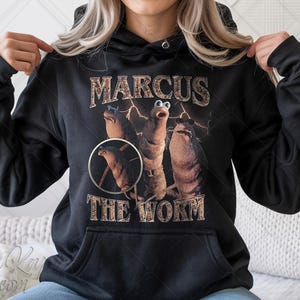 Pode incluir: Moletom preto com a frase "MARCUS THE WORM" em uma fonte de estilo vintage. O gráfico inclui três personagens semelhantes a vermes com raios. O moletom tem um bolso frontal e um capuz com cordão.