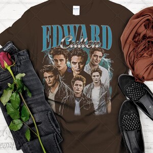 May include: Brown t-shirt featuring a graphic of Edward Cullen with the text "EDWARD Cullen" in blue and silver. The shirt is styled with black jeans, a rose, a brown scarf, and black loafers.