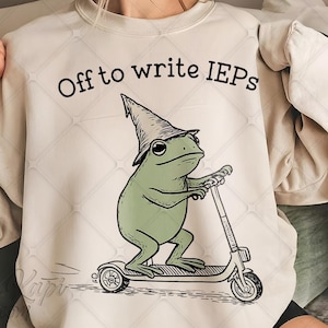 May include: Cream-colored sweatshirt with the text "Off to write IEPs." The design features a green frog wearing a wizard hat and riding a scooter. The frog is the central focus of the graphic.