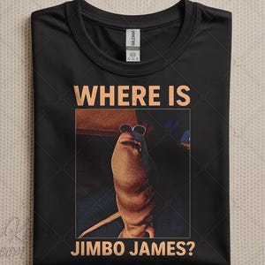 Pode incluir: Camiseta preta com o texto "WHERE IS JIMBO JAMES?" em cor de cobre. A camiseta apresenta uma imagem emoldurada de um fantoche usando óculos de sol. O design é centralizado na camiseta.