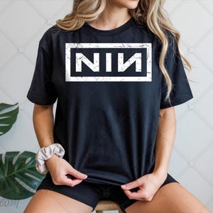 May include: Black t-shirt featuring the letters "NIN" in a white, distressed rectangular design. The short-sleeved top is made of a soft fabric. The style is casual and relaxed, suitable for everyday wear.