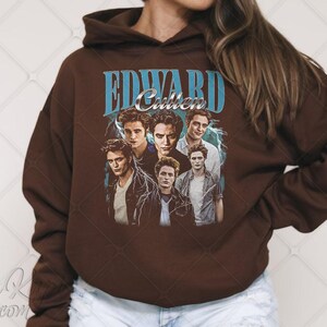 May include: A brown hooded sweatshirt featuring a graphic of Edward Cullen from the Twilight series. The design includes the name "EDWARD Cullen" in blue text with images of the character. The sweatshirt is a solid brown color.