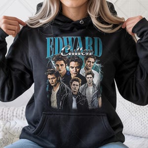 May include: Black hooded sweatshirt featuring a graphic of Edward Cullen from the Twilight series. The design includes the name "Edward Cullen" in teal lettering above a collage of images of the character. The sweatshirt has a front pocket.