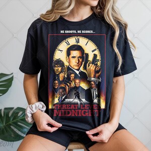 May include: Black t-shirt with a movie poster design featuring the text "HE SHOOTS, HE SCORES..." and "THREAT LEVEL MIDNIGHT". The design includes a clock face and several characters. The shirt is worn by a person.