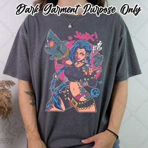 May include: Dark gray t-shirt featuring a colorful graphic of a female character with blue hair, holding a weapon. The design includes the word "Jinx" and other text. The shirt is a dark garment.