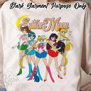 May include: A cream-colored t-shirt featuring a graphic print of the Sailor Moon characters and the text "Sailor Moon" in a stylized yellow and purple font. The design includes five anime figures in colorful outfits and a small blue cat.
