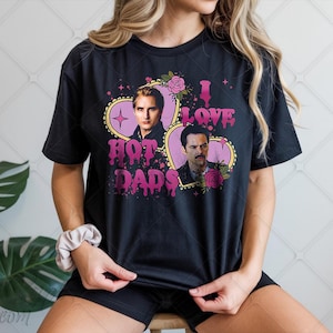 Puede incluir: Camiseta negra con un diseño gráfico que presenta dos retratos dentro de marcos en forma de corazón. El texto "I Love Hot Dads" está en rosa, con un efecto de goteo. Acentos florales y estrellas completan el diseño. La camiseta la lleva una persona.