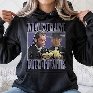 Może przedstawiać: Czarna bluza z kapturem z grafiką przedstawiającą dwóch mężczyzn i napisem "WHAT EXCELLENT BOILED POTATOES" w fioletowym pudełku. Projekt jest ilustracją w stylu vintage. Bluza ma przednią kieszeń i kaptur ze sznurkiem.