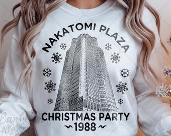 May include: White sweatshirt featuring a black graphic of the Nakatomi Plaza building, snowflakes, and text that reads "Nakatomi Plaza Christmas Party 1988". The design is centered on the front of the sweatshirt.