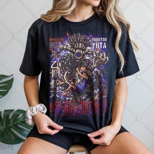 Peut inclure: T-shirt noir avec un graphisme représentant deux personnages s'embrassant, avec un grand monstre derrière eux. Le texte "OKKOTSU YUTA" est au-dessus des personnages, avec du texte japonais en dessous. Le design est dans les tons de violet, noir et gris.