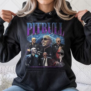 Puede incluir: Sudadera con capucha negra con un gráfico de Pitbull en varias poses, y el nombre "PITBULL" en un diseño púrpura y azul. El diseño tiene una estética vintage y retro.