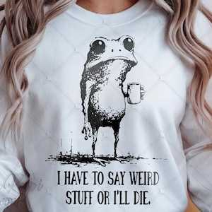 Könnte beinhalten: Weißes Sweatshirt mit einer schwarzen Illustration einer Froschfigur, die eine Tasse hält. Der Frosch hat einen mürrischen Gesichtsausdruck. Darunter steht der Text: "I HAVE TO SAY WEIRD STUFF OR I'LL DIE".