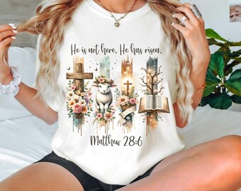 May include: White t-shirt featuring a watercolour design of crosses, flowers, a lamb, and an open book. The text "He is not here. He has risen. Matthew 28:6" is printed on the shirt.