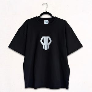 May include: A black short-sleeve t-shirt featuring a centered, stylized white graphic of a skull-like mask. The shirt has a crew neckline and is displayed on a black hanger. The graphic design is positioned on the chest area of the garment.