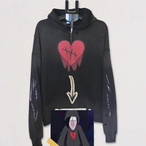 May include: A black zip-up hoodie featuring a red heart graphic with black stitching details on the chest, a beige downward-pointing arrow below, and white stitch patterns on the sleeves. A small square image of a cartoon character is attached.