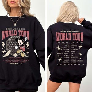 Könnte beinhalten: Schwarzes Sweatshirt mit dem Text "DRINK AROUND THE WORLD TOUR" in Rosa. Mit einer Mickey Mouse-Grafik und einer Liste von Städten. Auf der Rückseite des Sweatshirts steht ebenfalls "DRINK AROUND THE WORLD TOUR" und eine Liste von Städten.