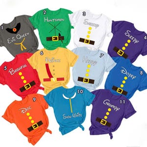 Könnte beinhalten: Eine Sammlung von elf farbenfrohen T-Shirts mit Designs zum Thema Charaktere. Jedes Shirt hat eine andere Farbe und ein einzigartiges Designelement, einschließlich Text wie "Evil Queen", "Huntsman", "Sneezy", "Sleepy", "Bashful", "Prince", "Happy", "Dopey", "Doc", "Snow White" und "Grumpy".